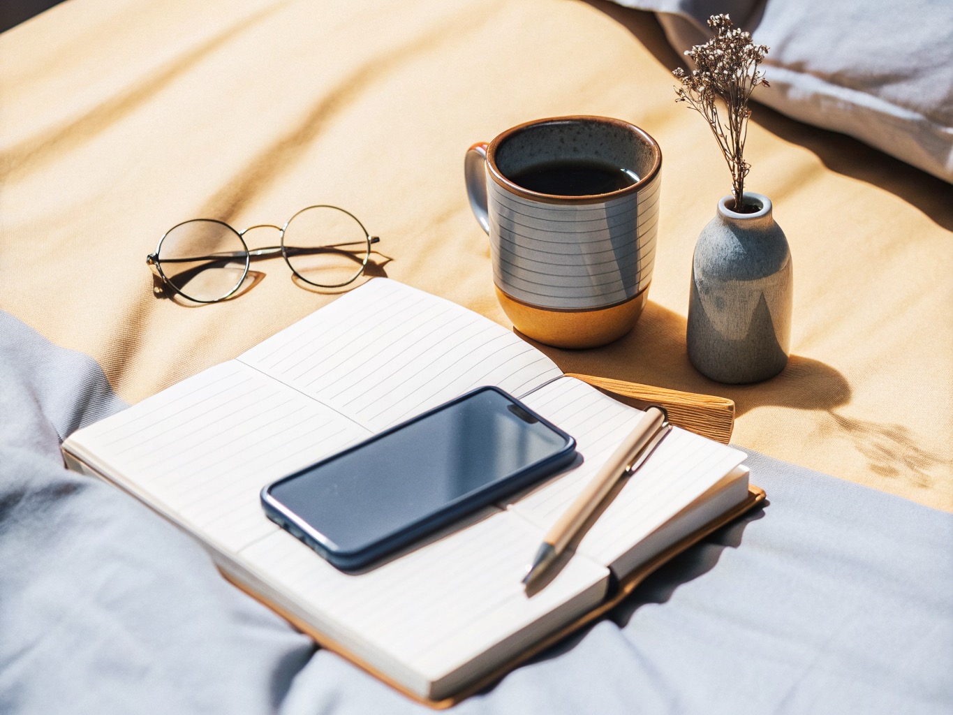 A phone resting on an open notebook beside reading glasses, a cup of coffee, and a small bud vase in warm natural light